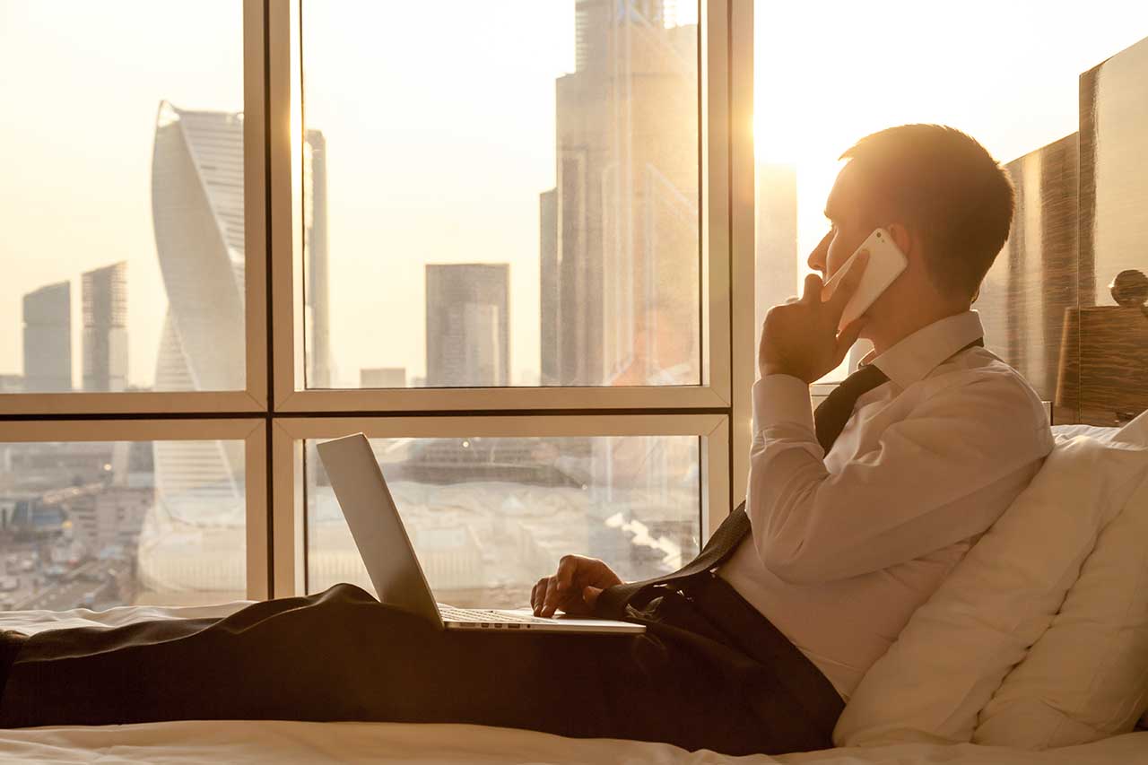 Businessman sitting on the bed with laptop in modern room and looking at sunny city view in window
